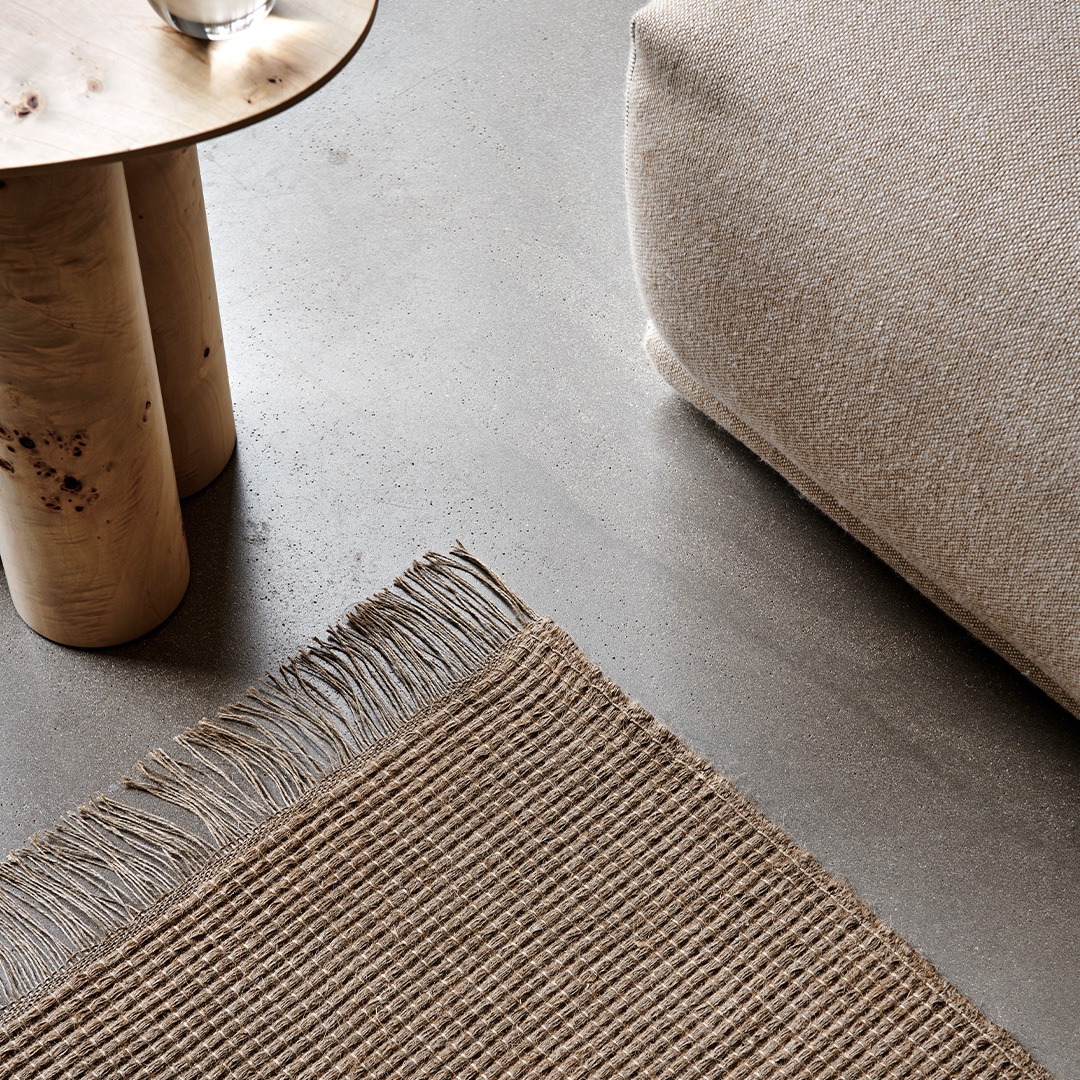 A close-up of a linen woven rug with fringes on a smooth, gray floor, next to a wooden table and a beige upholstered piece of furniture.